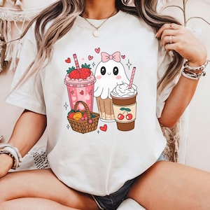 May include: White t-shirt featuring a graphic of a ghost wearing a pink bow, holding a milkshake, a cupcake, and a coffee cup. The design also includes hearts, strawberries, and a basket of fruit.