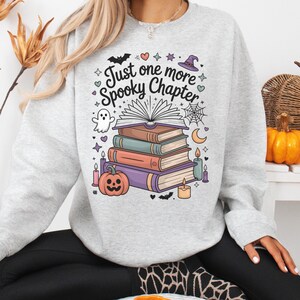May include: Light gray sweatshirt with a colorful graphic featuring a stack of books, a pumpkin, a ghost, and the text "Just one more Spooky Chapter." The design includes bats, stars, and a witch's hat, perfect for Halloween or book lovers.