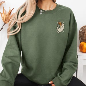 May include: An olive green sweatshirt featuring a small embroidered graphic of a cat holding a slice of pizza. The cat is light beige with a white belly and is surrounded by small white stars. The sweatshirt has a crew neck and long sleeves.