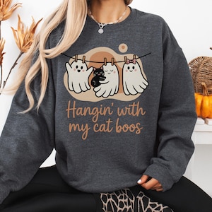 Könnte beinhalten: Dunkelgraues Sweatshirt mit einem Halloween-Motiv. Das Design zeigt drei Cartoon-Geister auf einer Wäscheleine mit einer schwarzen Katze. Der Text lautet "Hangin' with my cat boos".