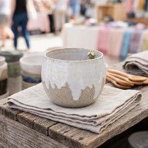 Può includere: Una tazza in ceramica rotonda con base beige maculata e finitura a smalto bianco colante. Una piccola figura tridimensionale di un'ape gialla e nera è fissata sul bordo interno della tazza. La tazza poggia su un tovagliolo di lino beige chiaro.