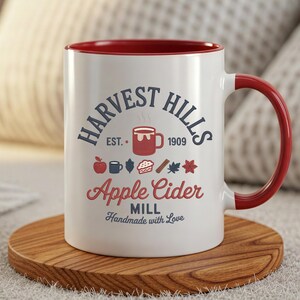 May include: White ceramic mug with a red interior and handle, featuring the text "HARVEST HILLS APPLE CIDER MILL" and related graphics. The mug sits on a round wooden coaster.