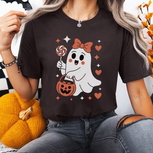 May include: A dark brown t-shirt featuring a cartoon ghost holding a lollipop and a pumpkin-shaped basket. The ghost has a bow and is surrounded by stars and hearts. A fun Halloween-themed graphic tee.