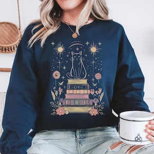 May include: A navy blue sweatshirt featuring a graphic of a black cat perched atop a stack of books. The cat is surrounded by stars, flowers, and a crescent moon. The text "I LOVE BOOKS" is printed on the books.