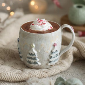 Könnte beinhalten: Ein Keramikbecher mit gesprenkelter, cremeweißer Glasur und geprägten, handbemalten grünen und weißen Tannenbaummotiven. Der Becher ist mit dunkelbrauner heißer Schokolade gefüllt, die mit weißer Schlagsahne und zerstoßenen Pfefferminzstücken garniert ist.