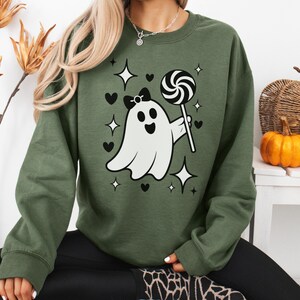 Puede incluir: Una sudadera verde oliva con un fantasma de dibujos animados sosteniendo un caramelo negro y blanco. El fantasma tiene un lazo negro y está rodeado de corazones negros y formas de estrella. La sudadera es parte de una colección temática de Halloween.