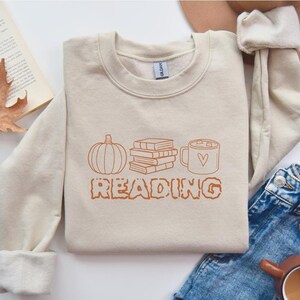 May include: A beige sweatshirt with a brown graphic of a pumpkin, a stack of books, a mug of hot chocolate, and the word "READING" in a stylized font.