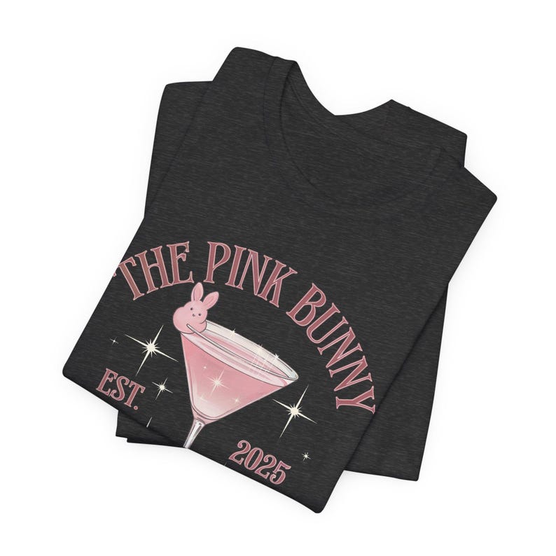 A trendy cocktail-themed t-shirt featuring a pink martini glass with a bunny accent and vintage lettering, perfect for bar lovers and aesthetic outfits. The main colors are pink and white.
