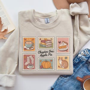 May include: Beige sweatshirt with a graphic of six postage stamp-style images. The images include a cup of coffee, books, a slice of pie, socks, a pumpkin, and a cat. The text "Chapter One: Apple Pie" is in the center.