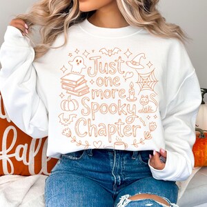 May include: White sweatshirt with the text "Just one more Spooky Chapter" in orange. The design includes a ghost, books, a pumpkin, bats, a witch hat, a spiderweb, a candle, a cat, and flowers. A cozy fall-themed top.