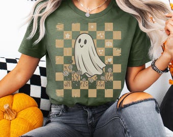 Pode incluir: Camiseta verde-oliva com um desenho de fantasma sorridente em um fundo xadrez com flores e outros pequenos gráficos. O fantasma é branco e o padrão xadrez é marrom e verde-oliva. A camiseta tem mangas curtas.