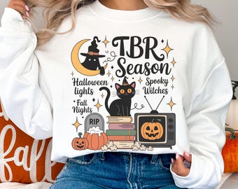 May include: White sweatshirt with a Halloween-themed graphic. The design includes a black cat wearing a witch hat, pumpkins, a tombstone, and the text "TBR Season". Additional text includes "Halloween lights", "Spooky Witches", and "Fall Nights".
