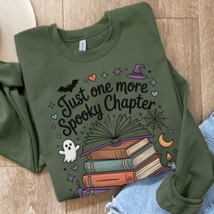May include: Olive green sweatshirt with the text "Just one more Spooky Chapter" and Halloween-themed illustrations. The design includes a stack of books, a ghost, a witch hat, and a spiderweb.