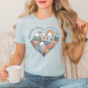 T-shirt featuring a colorful heart-shaped mountain design filled with wildflowers, sun, birds, and nature elements. Text reads “Adventure Awaits.” A cheerful outdoorsy aesthetic ideal for travel lovers, hikers, and boho nature girls.