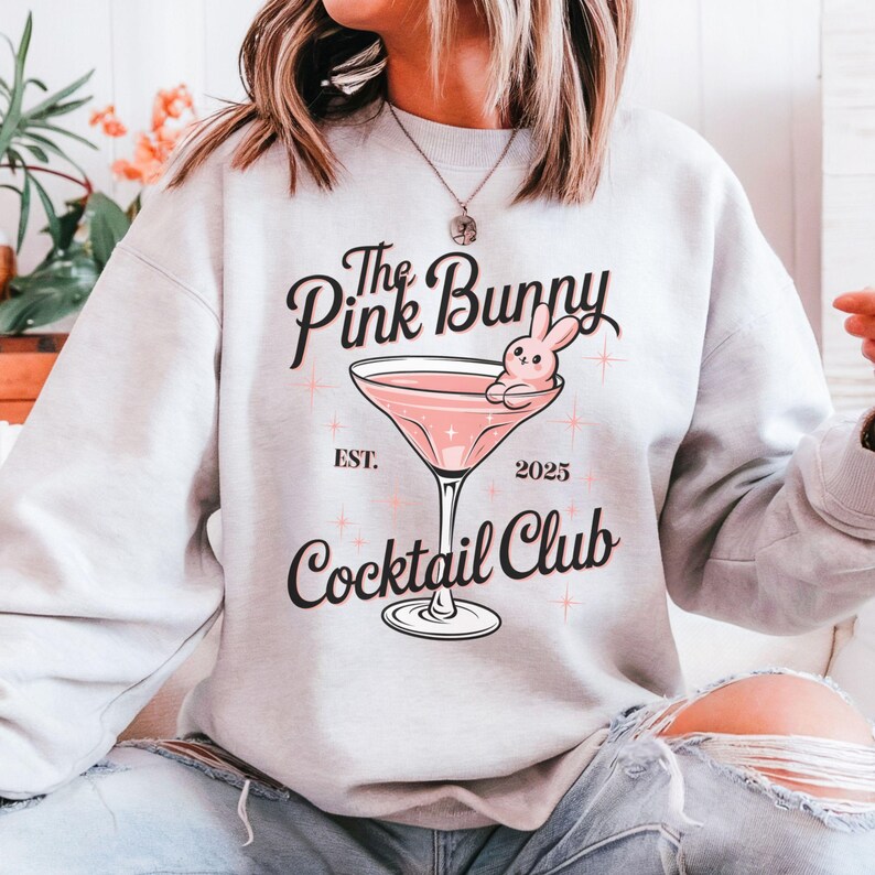 A sweatshirt featuring a pink martini glass with a bunny lounging on the rim, surrounded by vintage-style lettering that reads: The Pink Bunny Cocktail Club, Est. 2025. The main colors are pink and black.