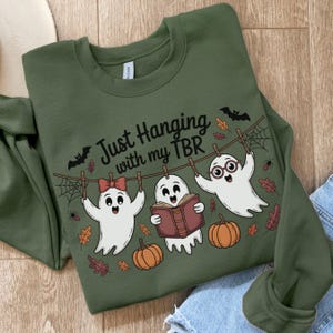 May include: Olive green sweatshirt with the text "Just Hanging with my TBR." The graphic features three cartoon ghosts hanging on a clothesline with pumpkins, bats, and fall leaves. One ghost wears glasses and another a bow.
