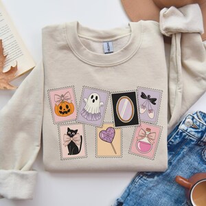 May include: Beige sweatshirt featuring a collection of whimsical Halloween-themed postage stamp designs. The stamps showcase a pumpkin, ghost, mirror, ballet shoes, black cat, heart lollipop, and potion bottle, all with a vintage aesthetic.