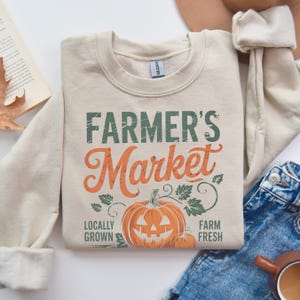 Może przedstawiać: Beżowa bluza z napisem "FARMER'S Market" w zielonych i pomarańczowych literach. Bluza ma grafikę dyni z napisem "LOCALLY GROWN" i "FARM FRESH" poniżej.
