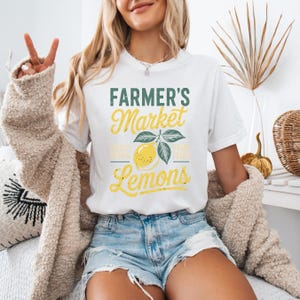 T-shirt with vintage-style “Farmer’s Market Lemons” graphic featuring a yellow lemon with leaves and rustic text. The main colors are yellow and green.