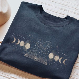 Puede incluir: Sudadera azul marino con una ilustración dorada de un sombrero de bruja, un libro abierto y fases lunares. El diseño incluye estrellas, creando un tema místico. La sudadera está hecha de un material suave.