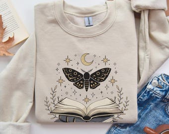 May include: Beige sweatshirt featuring a black moth design with a crescent moon, stars, and an open book. The graphic is surrounded by botanical elements. The sweatshirt is made of a soft material and is suitable for casual wear.