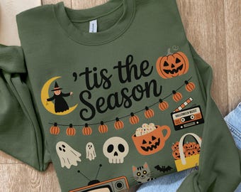 May include: Olive green sweatshirt with a Halloween-themed graphic. The design includes a witch on a crescent moon, pumpkins, ghosts, a skull, a cat, and the phrase "'tis the Season". A cassette tape with "Halloween Music" is also featured.