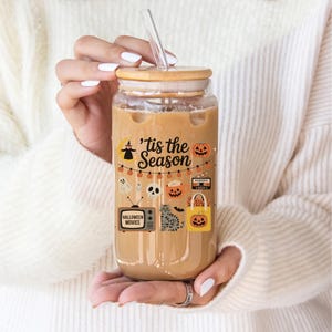 Könnte beinhalten: Ein klares Glas mit einem kaffee farbigen Getränk, mit einem Bambusdeckel und einem durchsichtigen Strohhalm. Das Glas ist mit Halloween-Aufklebern verziert, darunter Kürbisse, Geister und der Text "'tis the Season".