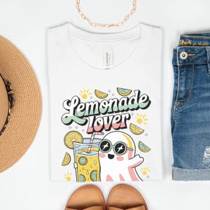 Peut inclure: T-shirt blanc avec l'inscription "Lemonade Lover" dans une police r&eacute;tro, repr&eacute;sentant un fant&ocirc;me de dessin anim&eacute; portant des lunettes de soleil et tenant un verre de limonade. Le motif comprend des tranches de citron et des &eacute;clats de soleil.