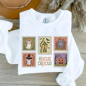 Könnte beinhalten: Weißes Sweatshirt mit sechs Illustrationen im Postkartenstil zum Thema Halloween. Designs umfassen einen Geist, Fledermäuse, eine Laterne, einen Grabstein, eine Eule und einen Kürbis. Die Worte "Hocus Crocus" sind in Grün und Orange gedruckt.