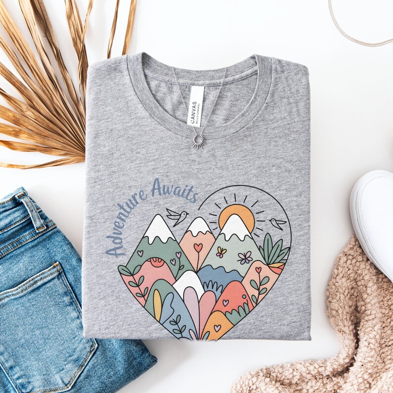 T-shirt featuring a colorful heart-shaped mountain design filled with wildflowers, sun, birds, and nature elements. Text reads “Adventure Awaits.” A cheerful outdoorsy aesthetic ideal for travel lovers, hikers, and boho nature girls.