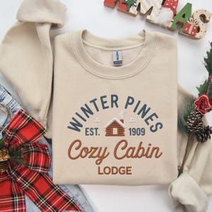 Pode incluir: Camisola bege com o texto "Winter Pines Cozy Cabin Lodge" em azul e castanho. O design inclui um gráfico de cabana e o ano "1909". Uma peça de vestuário com tema de inverno.
