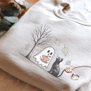 May include: Beige sweatshirt featuring a Halloween-themed illustration. A white ghost holding a pumpkin stands near a tree, with a black cat looking up. The design includes falling leaves, creating a cozy autumn scene. A gold necklace and ring are also visible.