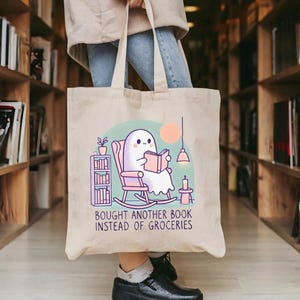A natural-colored tote bag featuring a cute ghost reading a book with a humorous message about choosing books over groceries. The text says: Bought Another Book Instead Of Groceries. Main colors are blue, orange, white, pink and black.