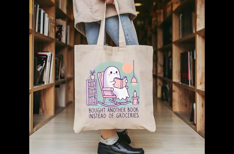 A natural-colored tote bag featuring a cute ghost reading a book with a humorous message about choosing books over groceries. The text says: Bought Another Book Instead Of Groceries. Main colors are blue, orange, white, pink and black.