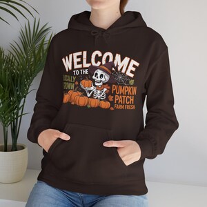 May include: A dark brown hoodie with a front pocket and a drawstring hood. The graphic on the front says "WELCOME TO THE PUMPKIN PATCH" with a skeleton holding pumpkins. The words "Locally Grown" and "Farm Fresh" are also included.