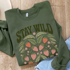 May include: A green sweatshirt with the text "Stay Wild" in a retro font. The text is surrounded by a design of green leaves and pink strawberries.