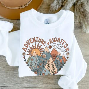 Könnte beinhalten: Weißes Sweatshirt mit einem farbenfrohen Grafikdesign. Das Design zeigt eine herzförmige Illustration mit Bergen, einer Sonne, Blumen und den Worten "Adventure Awaits". Das Sweatshirt hat einen Rundhalsausschnitt und lange Ärmel.