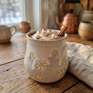 Op de afbeelding: Een gespikkelde keramieken mok met een crèmekleurig glazuur en reliëfpatronen van witte madeliefjes. De mok is gevuld met warme chocolademelk, afgewerkt met slagroom, witte marshmallows, een snufje cacaopoeder en twee kaneelstokjes.