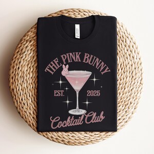 A trendy cocktail-themed t-shirt featuring a pink martini glass with a bunny accent and vintage lettering, perfect for bar lovers and aesthetic outfits. The main colors are pink and white.
