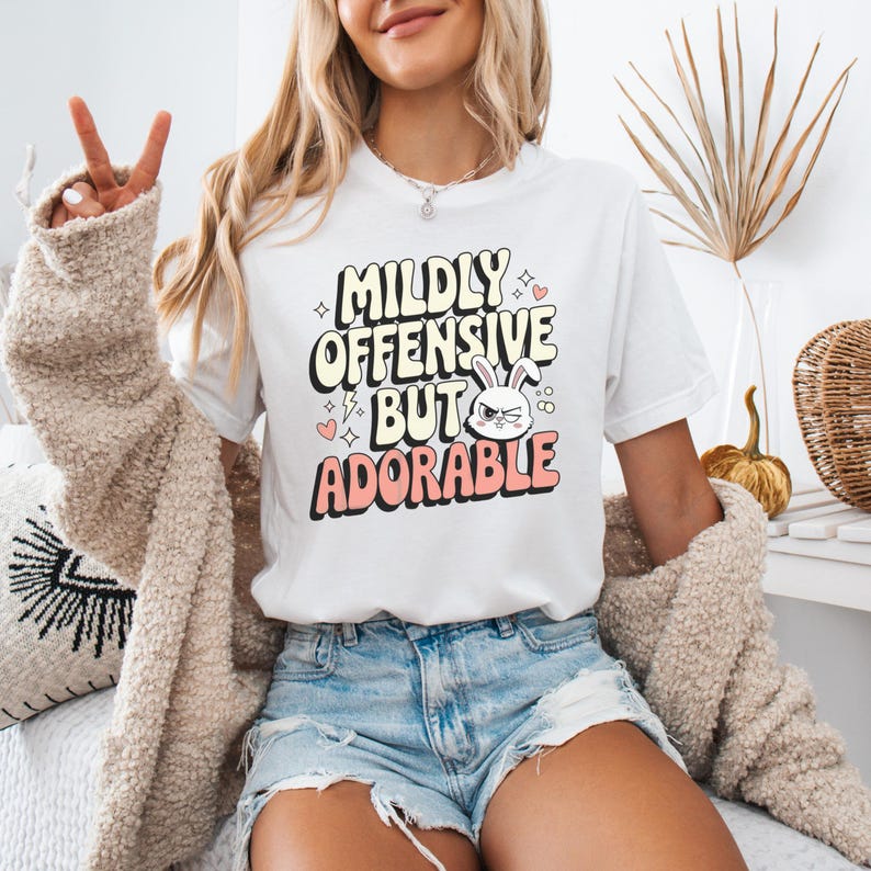 K&ouml;nnte beinhalten: Wei&szlig;es T-Shirt mit einer pinkfarbenen und wei&szlig;en Grafik, auf der "Mildly Offensive But Adorable" steht, mit einem Comic-Kaninchen mit w&uuml;tendem Gesichtsausdruck.