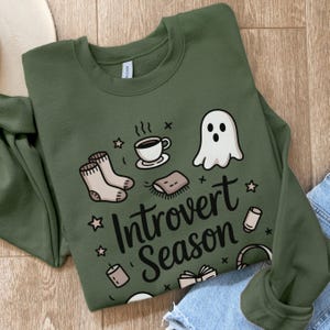 May include: Olive green sweatshirt with the words "Introvert Season" and illustrations of a coffee cup, socks, a ghost, a book, and other cozy items. The sweatshirt is made of a soft material.