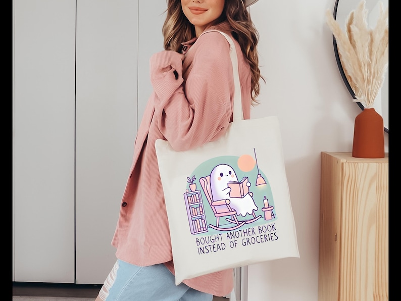 A natural-colored tote bag featuring a cute ghost reading a book with a humorous message about choosing books over groceries. The text says: Bought Another Book Instead Of Groceries. Main colors are blue, orange, white, pink and black.