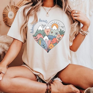 T-shirt featuring a colorful heart-shaped mountain design filled with wildflowers, sun, birds, and nature elements. Text reads “Adventure Awaits.” A cheerful outdoorsy aesthetic ideal for travel lovers, hikers, and boho nature girls.
