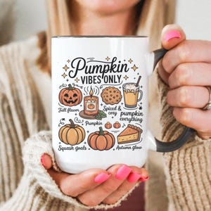May include: White ceramic mug with a black handle and rim, featuring the text "Pumpkin Vibes Only" and autumn-themed illustrations. The design includes pumpkins, a candle, a cookie, and a slice of pie. The mug is held by a person.