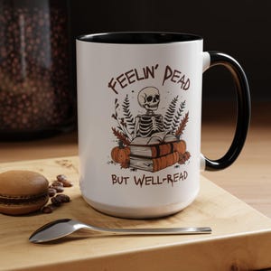 A white mug with a black or pink handle featuring a skeleton reading a book, surrounded by fall leaves and pumpkins, with the text: Feelin' Dead But Well-Read. Main colors are brown and orange.