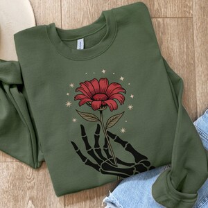 May include: An olive green sweatshirt featuring a red flower held by a black skeleton hand. The flower has a detailed design with a brown stem and is surrounded by small star-shaped accents. The sweatshirt is folded on a wooden surface.