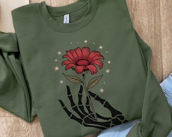 May include: An olive green sweatshirt with a red flower design held by a black skeleton hand. The flower has a detailed design with a brown stem and is surrounded by star-shaped accents. The sweatshirt is folded on a wooden surface.