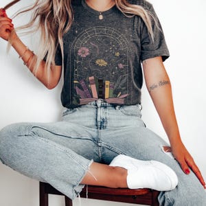 T-shirt featuring books, flowers, a snake, and stars framed by a mystical arched window—celestial, witchy, and dark academia styled.