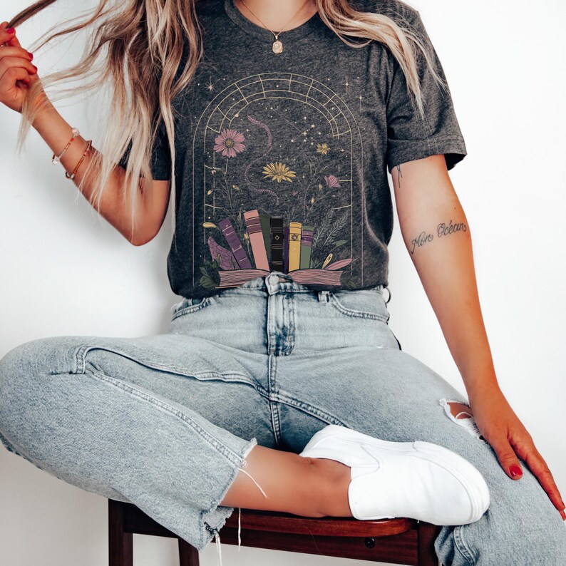 T-shirt featuring books, flowers, a snake, and stars framed by a mystical arched window—celestial, witchy, and dark academia styled.