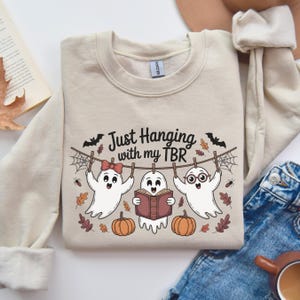 Op de afbeelding: Beige sweatshirt met de tekst "Just Hanging with my TBR". De afbeelding toont drie cartoongeesten die aan een waslijn hangen, met pompoenen en herfstbladeren. Eén geest draagt een bril en een ander een strik.